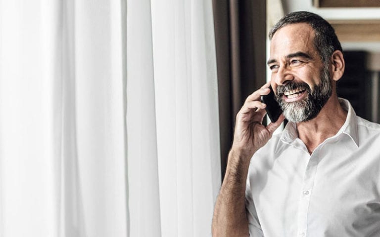 A smiling man with a white shirt speaking on a cell phone and looking out the window.