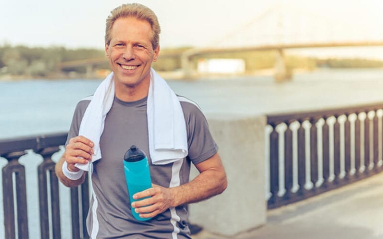 An athletic man holding a towel in one hand and water bottle in the other. The background is a riverfront with a bridge further in the background.