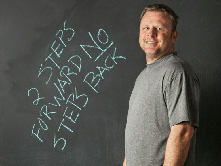 A man standing in front of a chalkboard. On the chalkboard is written "2 steps forward No Steps Back"