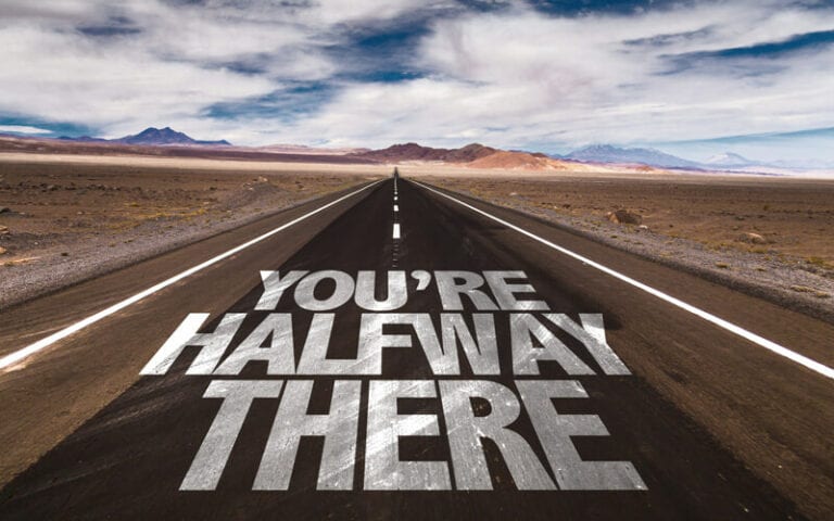 You're Halfway There is written on a desert road leading to some mountains in the distance.