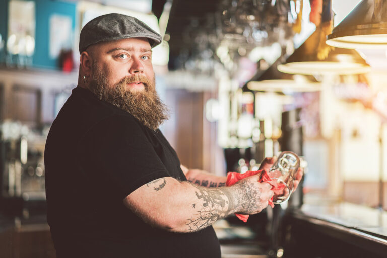 An overweight bearded bartender in a bar cleaning a glass.