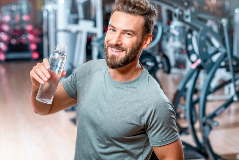 A smiling man wearing a silvery shirt in a gym setting holding an open bottle of water.
